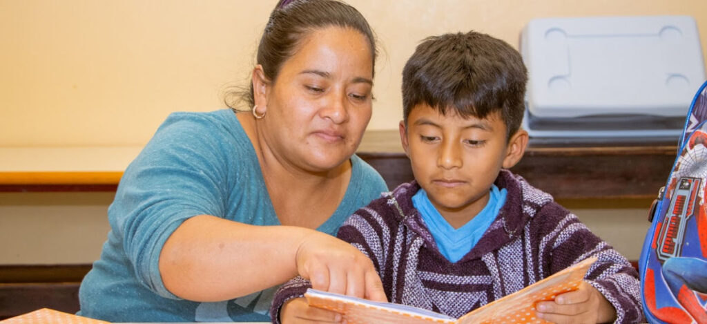 A guatemalan woman sitting beside a young boy and pointing at the words in a book he is reading