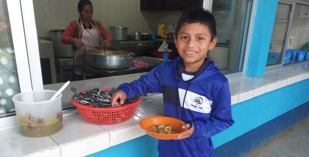 Boy in blue school uniform holding plate of food at cafeteria window. Woman cooking in the background.