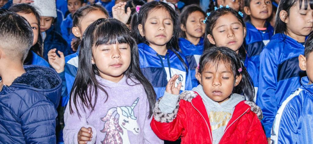 Young children, many in Impact School uniforms praying with eyes closed and hands raised