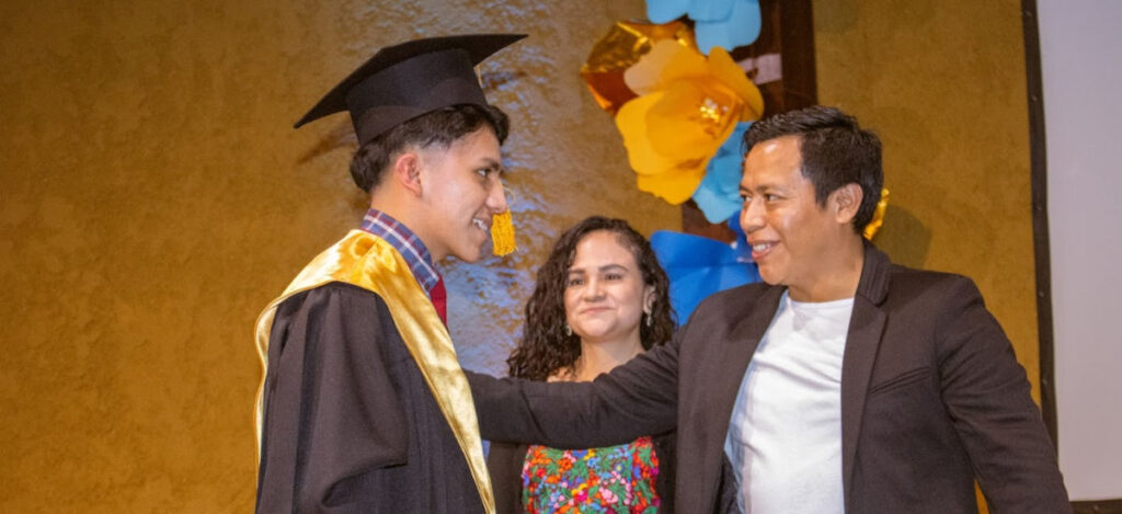 Pastor Hector congratulating a student in graduation gown while Ericka looks on